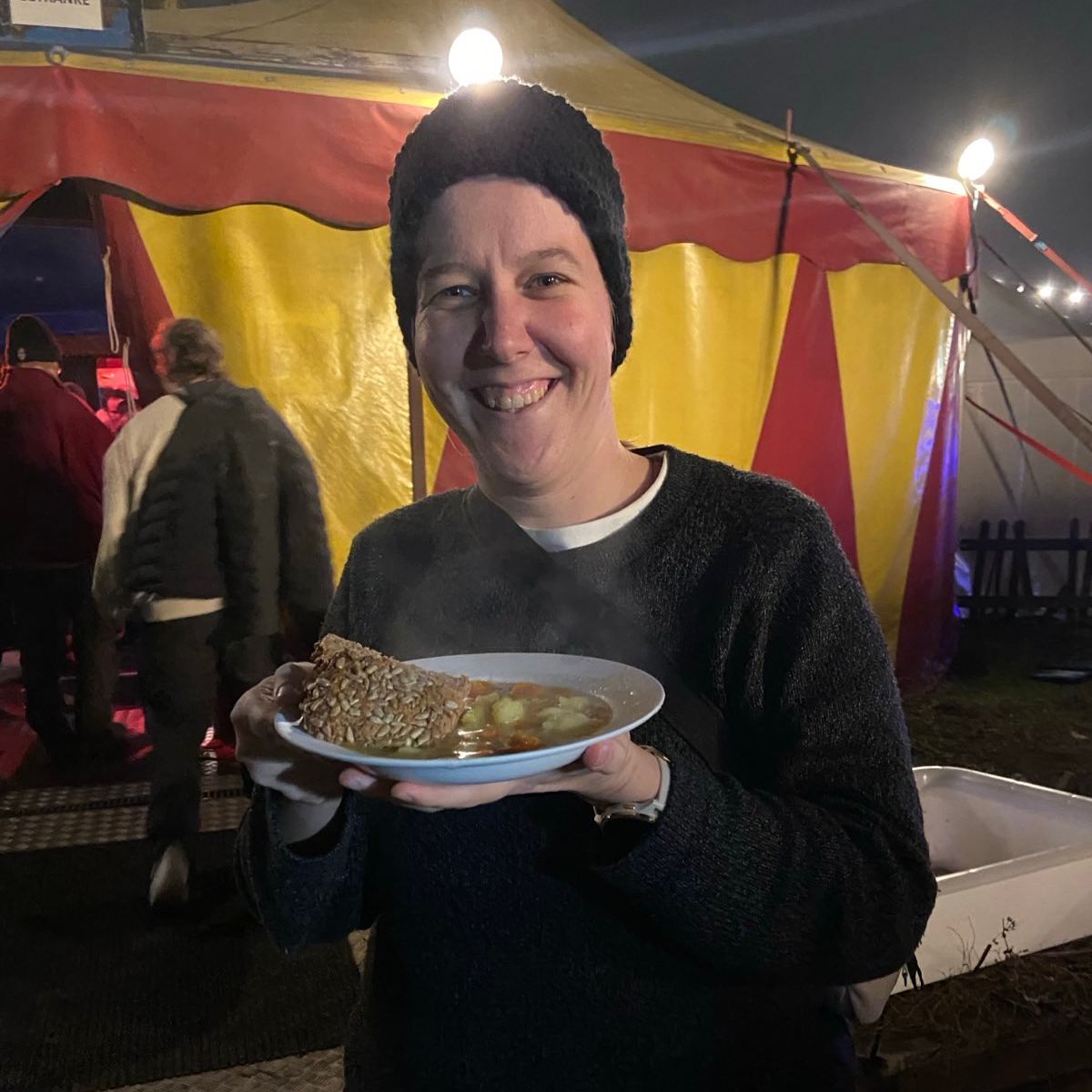 Frau mit Teller Suppe vor Zelt.
